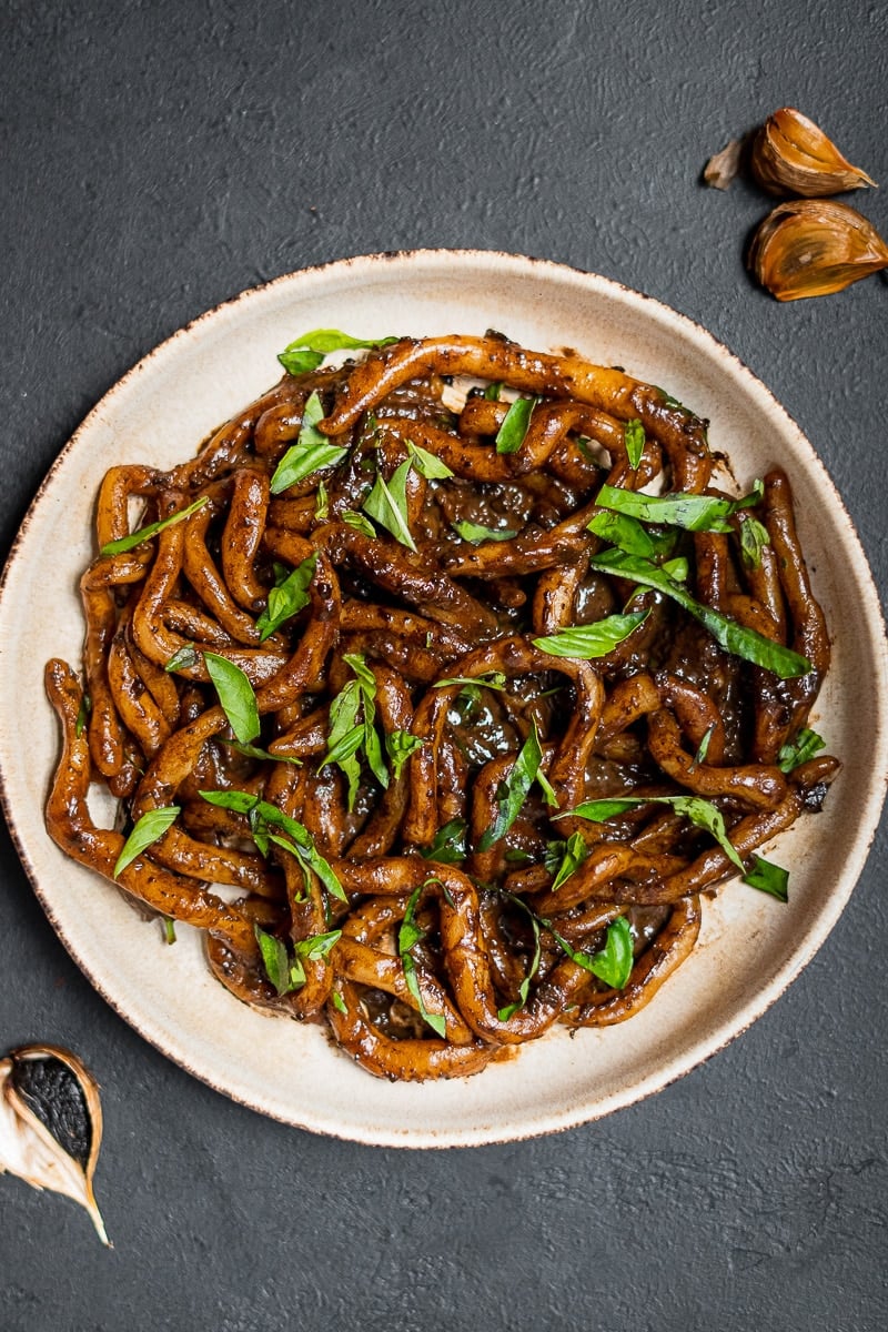 Black Garlic Pici on Black Background with Shredded Basil and black garlic cloves