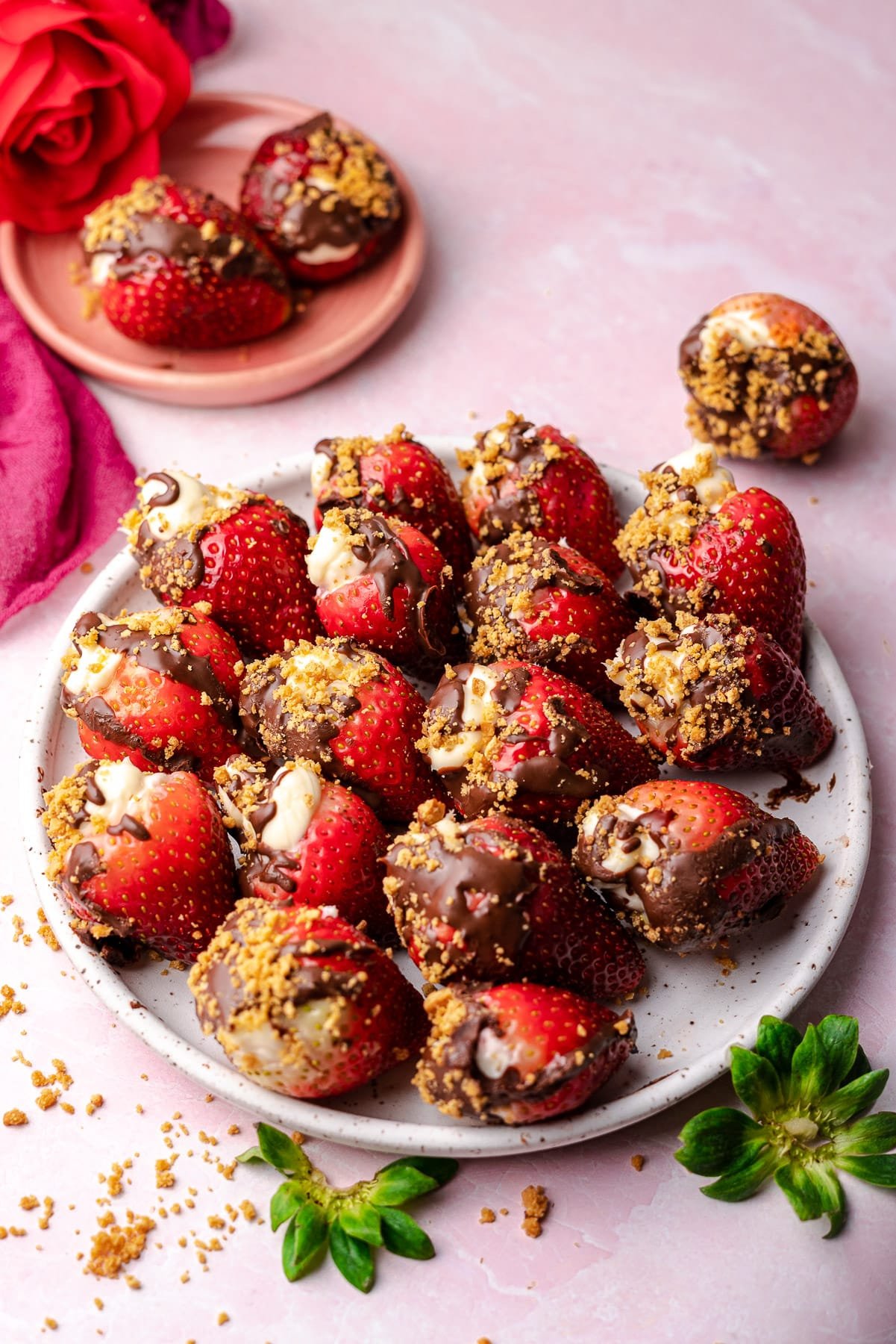 strawberries stuffed with cheesecake topped with chocolate and graham crackers in a plate with a rose