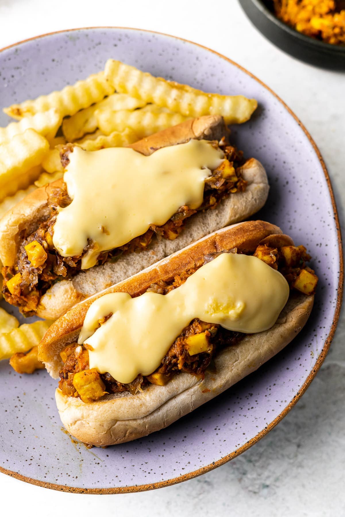 a close up at a 45 degree angle with paneer filling in a hot dog bun with cheese sauce and fries on the side