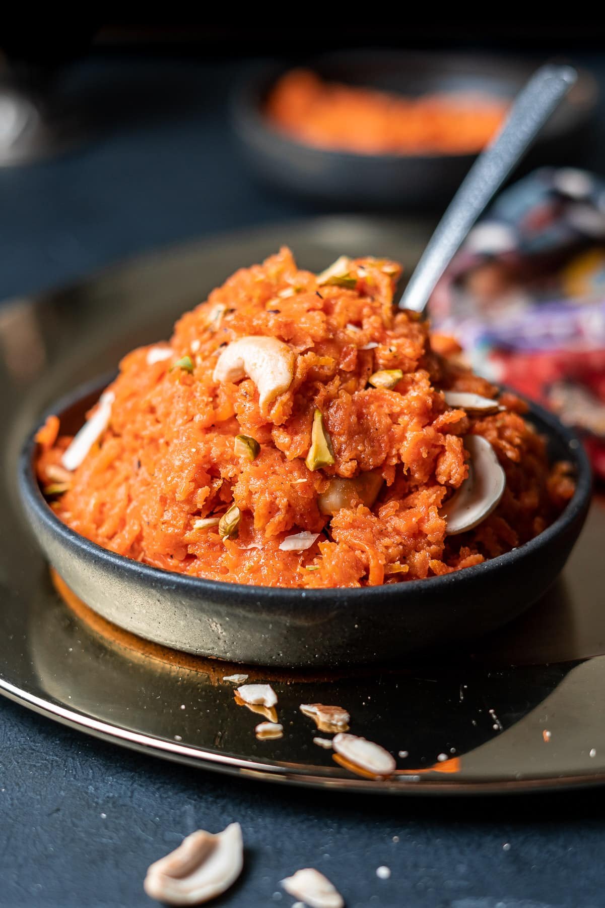 gajar ka halwa in a black bowl on a charger with a spoon sticking out of it and almond slivers