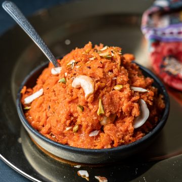 gajar ka halwa in a black bowl on a charger with a spoon sticking out of it and almond slivers