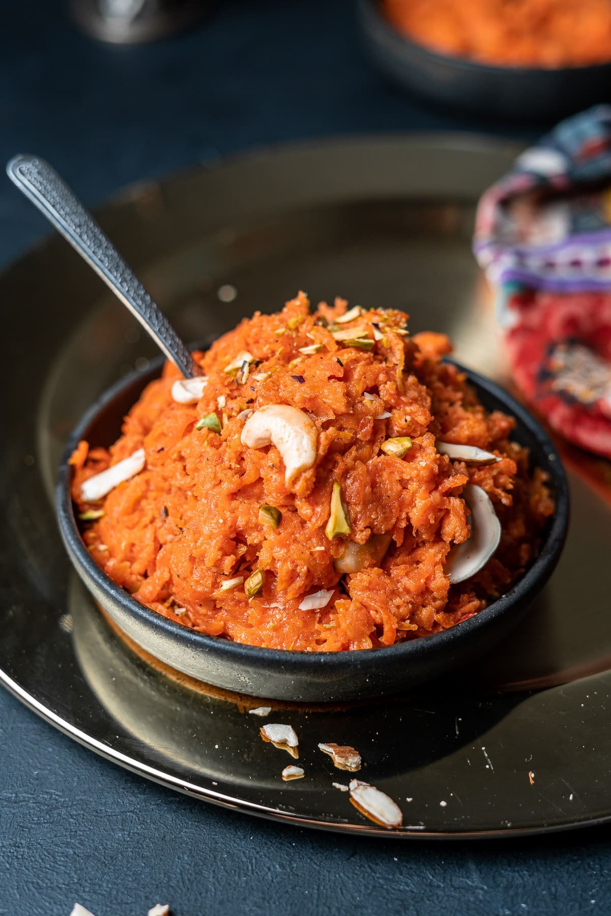 gajar ka halwa in a black bowl on a charger with a spoon sticking out of it and almond slivers