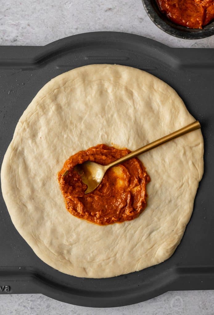 pizza dough on a brava tray with makhani sauce spread by a spoon