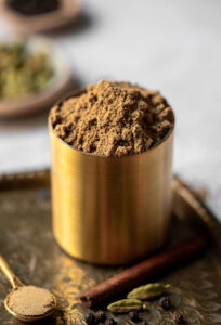 Ground Chai Spice in a Brass Container with whole spices in the foreground.