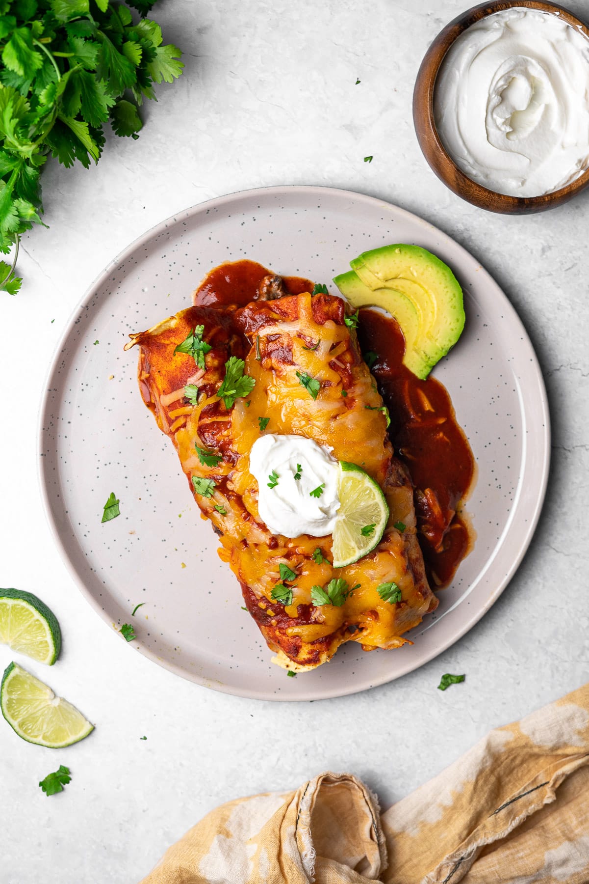 two enchiladas in a plate with sour cream, limes, cilantro, and avocado