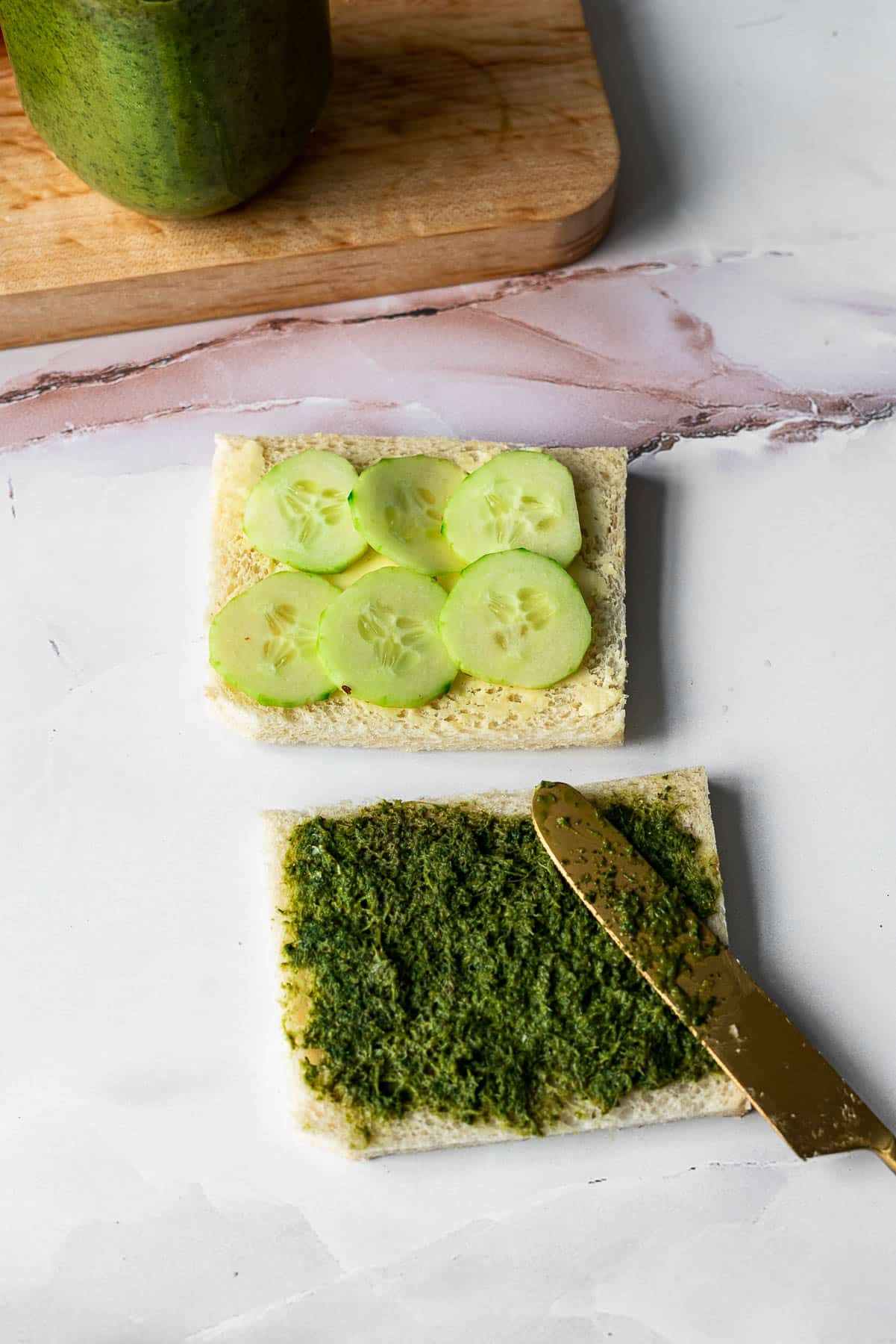 Crustless White bread with slices of cucumber on one and mint-cilantro chutney on the other with a golden knife resting on the chutney slice