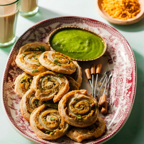 Samosa Pinwheels on a platter with appetizer picks and cilantro green chutney. Surrounded by other Indian snacks and chai.