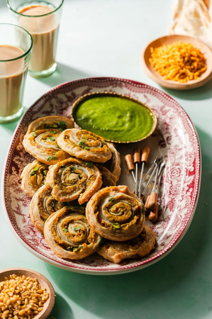 Samosa Pinwheels on a platter with appetizer picks and cilantro green chutney. Surrounded by other Indian snacks and chai.