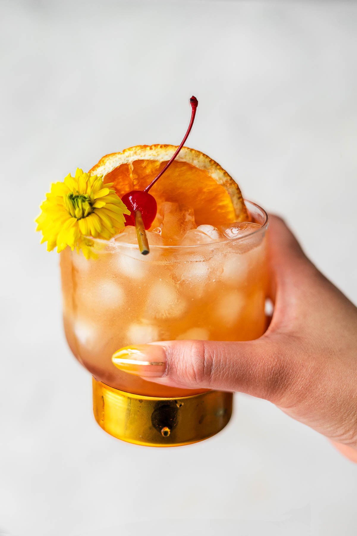 Manicured hand holding up a cup with amaretto sour with a gold bottom and cherry, flour, and orange slice as garnish