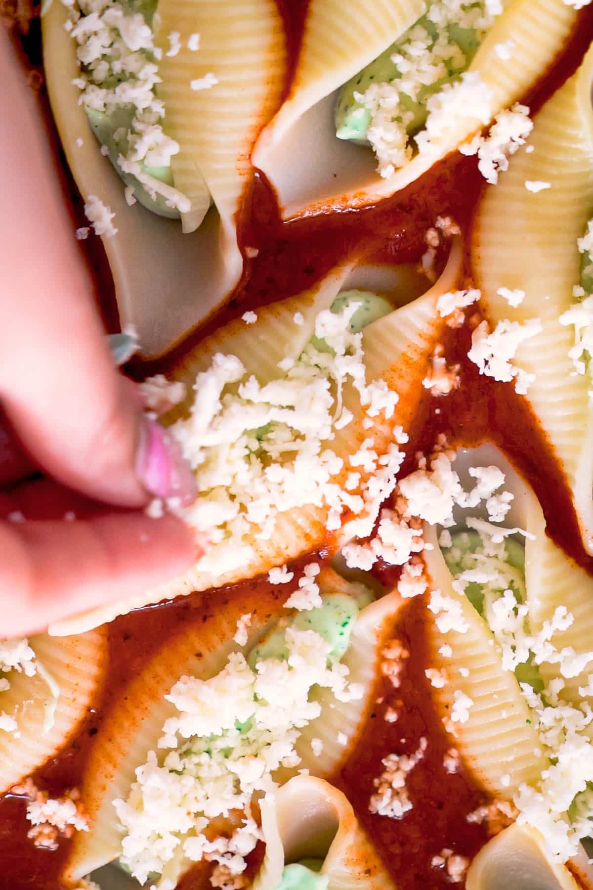 a hand spreading shredded mozzarella over jumbo shells filled with whipped ricotta set in tomato basil sauce