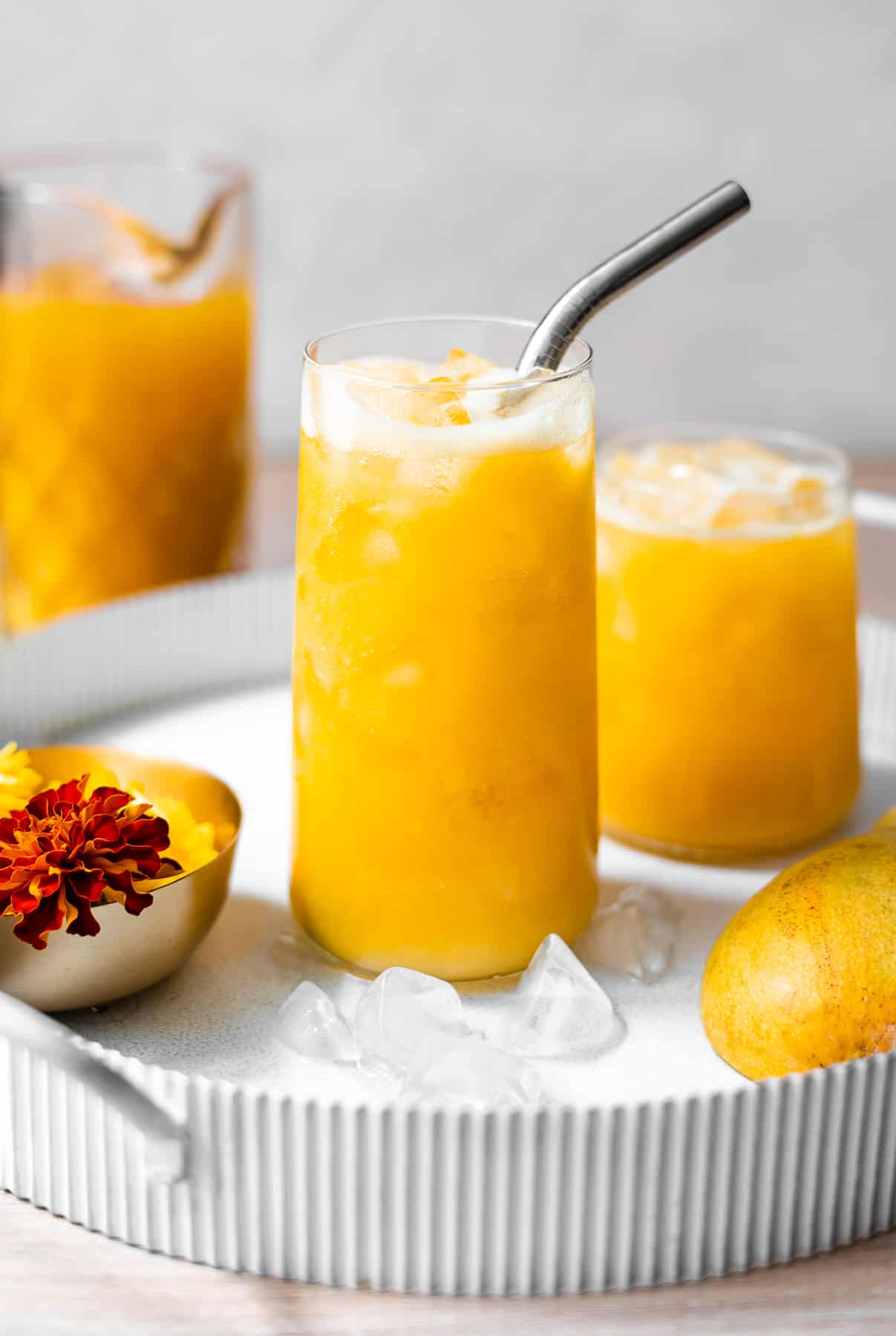 two sets of mango juice in glasses in a tray with flowers and fresh mangoes with a pitcher of mango juice