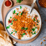Boondi Raita in a big bowl with a serving spoon on a platter with chili powder, cilantro, and boondi