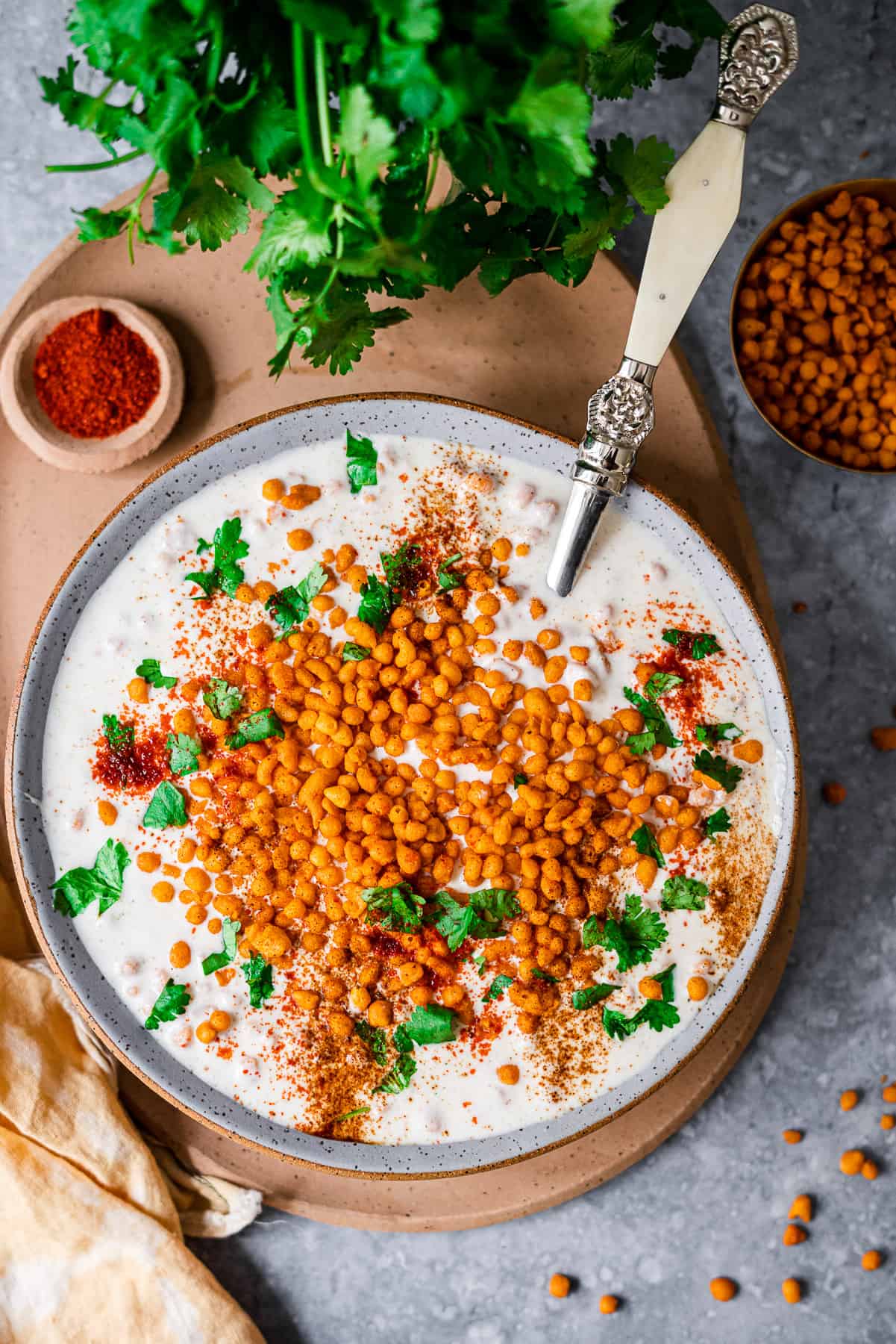 Boondi Raita in a big bowl with a serving spoon on a platter with chili powder, cilantro, and boondi