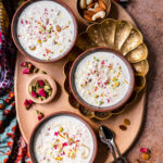 a platter with three bowls of kheer, nuts, raisins, roses, and cardamom