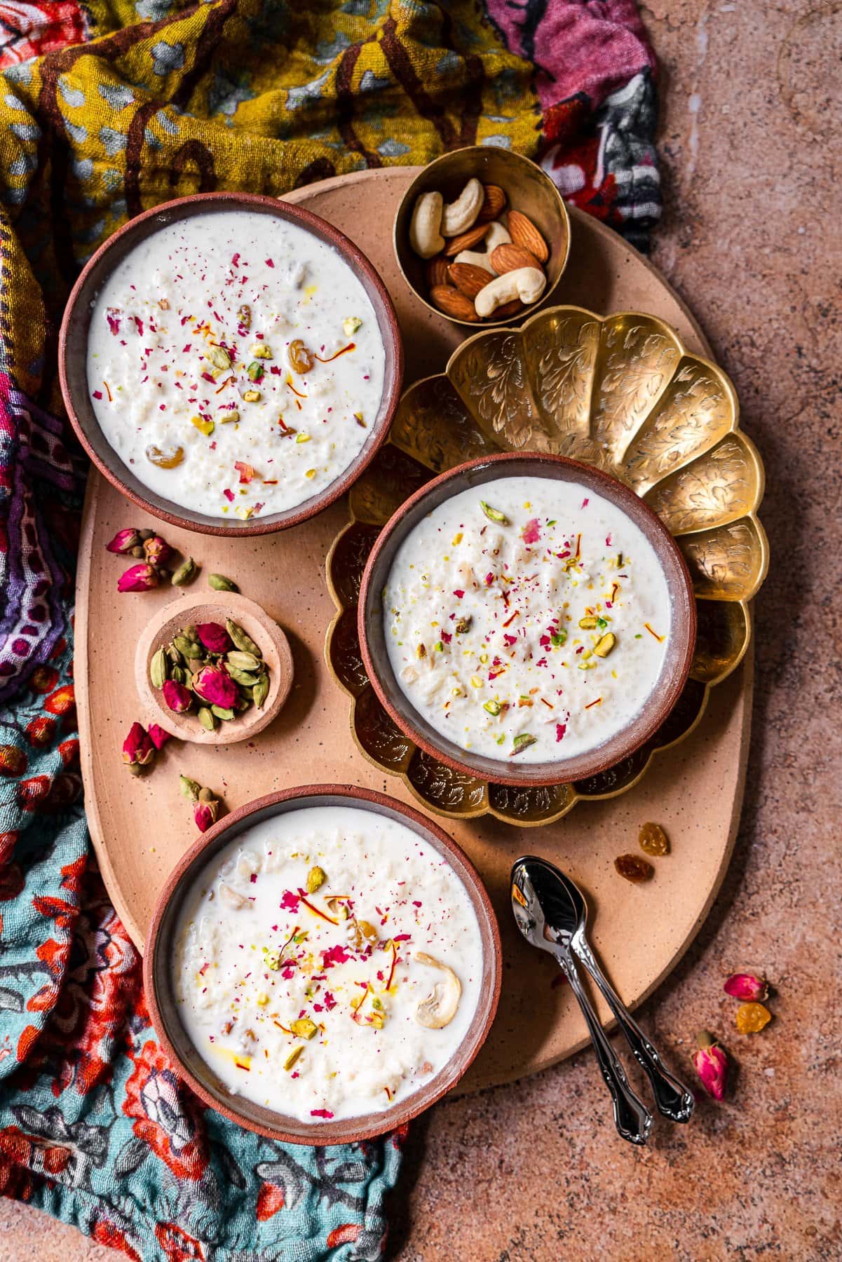 a platter with three bowls of kheer, nuts, raisins, roses, and cardamom