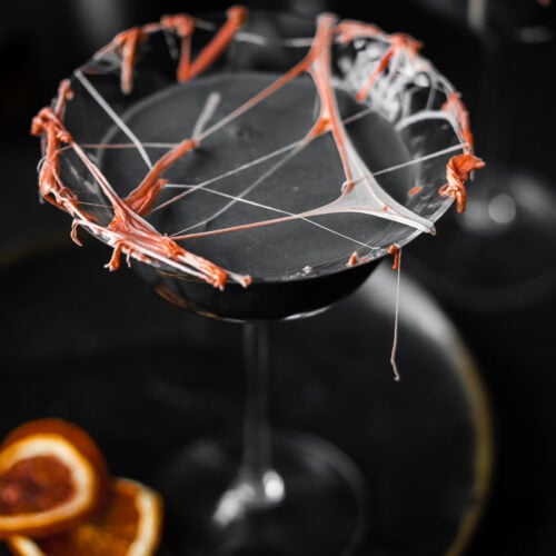 top shot of a coupe glass filled with a cocktail with marshmallow cob webs and dried blood oranges