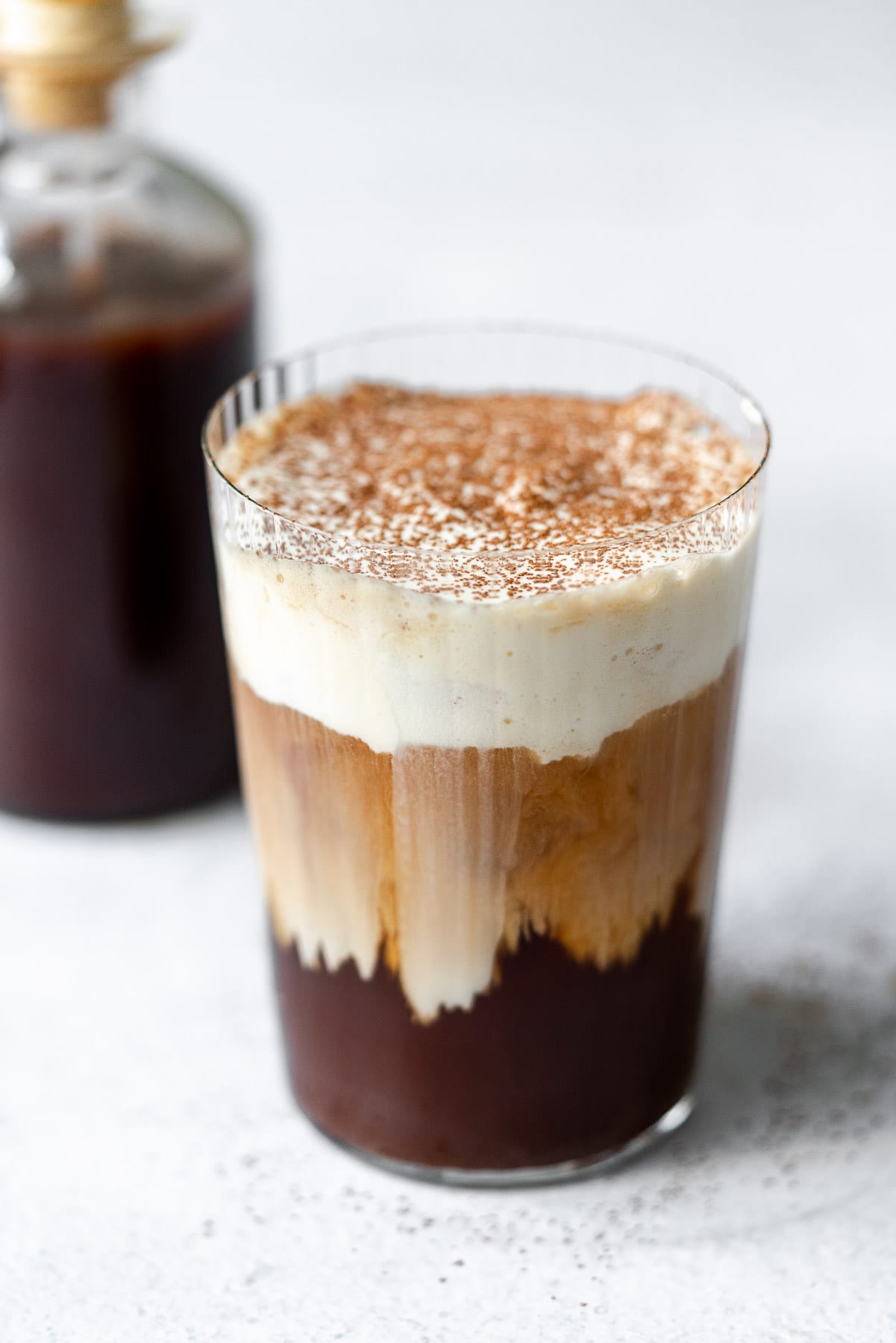 a glass filled with cold brew and sweet cream cold foam cascading down the glass topped with cocoa powder and a bottle of irish ceam syrup