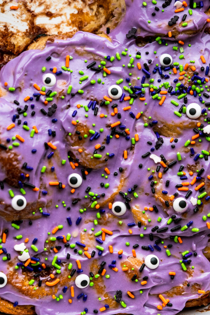 a baking dish full of cereal milk cinnamon rolls with ube cream cheese frosting with halloween sprinkles
