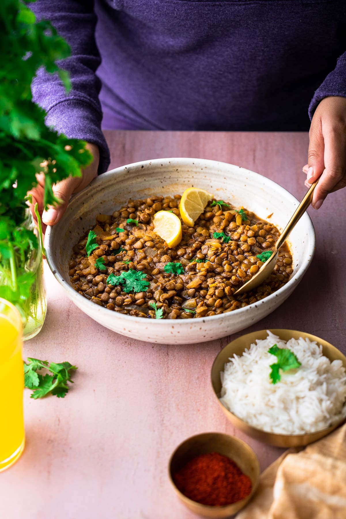 serving sabut masoor dal in a serving bowl with a person spooning it with rice, chili powder, cilantro, and juice alongside.