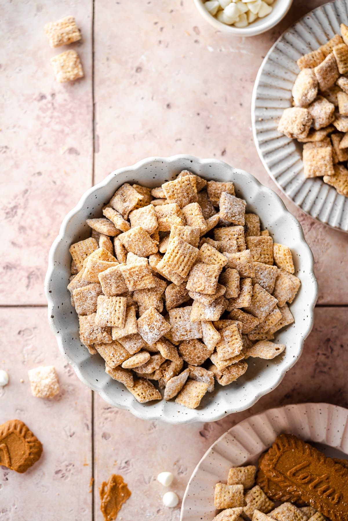 cookie butter puppy chow in a scalloped bowl with plates of puppy chow and biscoff