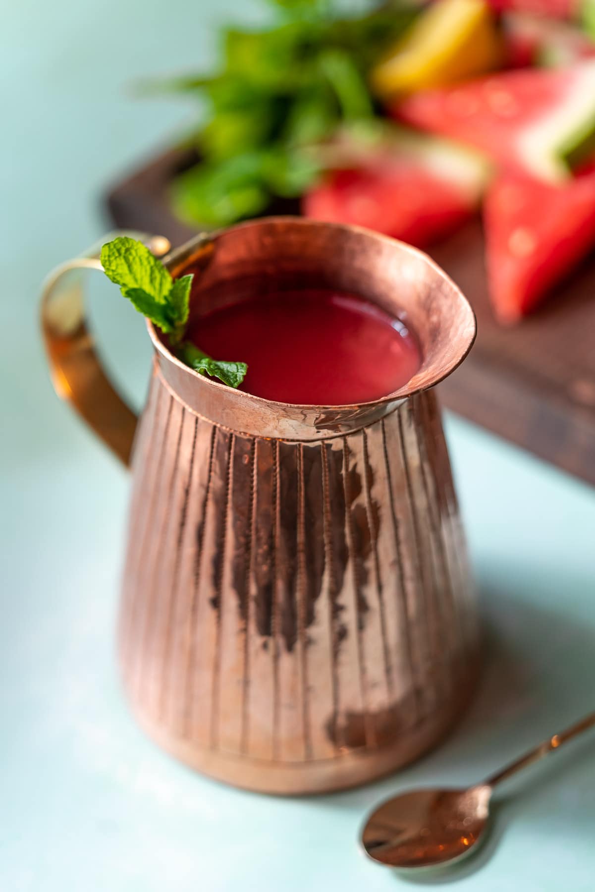 copper pitcher with watermelon rose cooler with mint and watermelon in the background on a cutting board