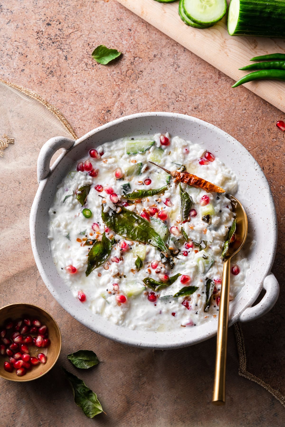 curd rice with pomegranates and curry leaves in a bowl with a gold spoon and fresh cucumbers and peppers on a board