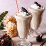 Two date shakes in milkshake glasses with whipped cream, a date, and a straw.