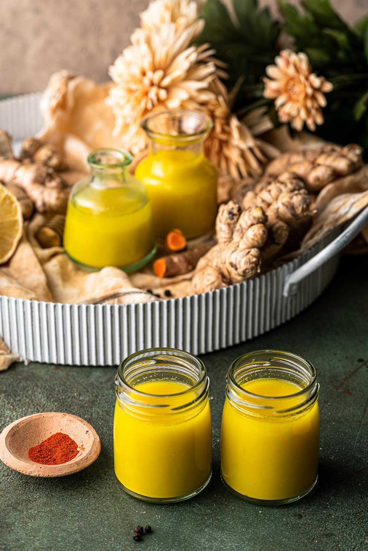 Four shots of ginger turmeric wellness shots with a tray full of the ingredients and flowers.