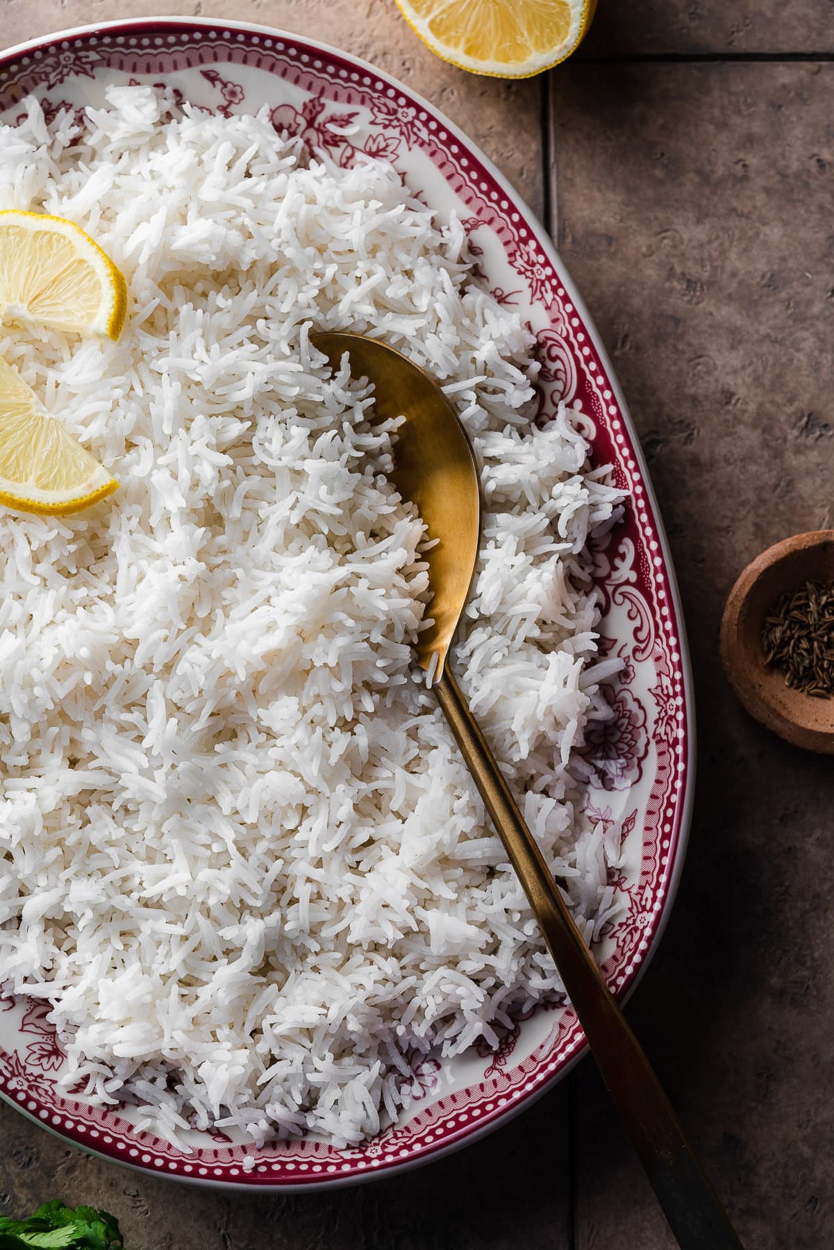 basmati rice in a decorative serving platter with lemon slices and a gold serving spoon with cumin seeds.