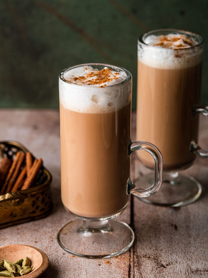 two irish coffee mugs with foamy chai tea lattes with whole cinnamon sticks and cardamom pods.