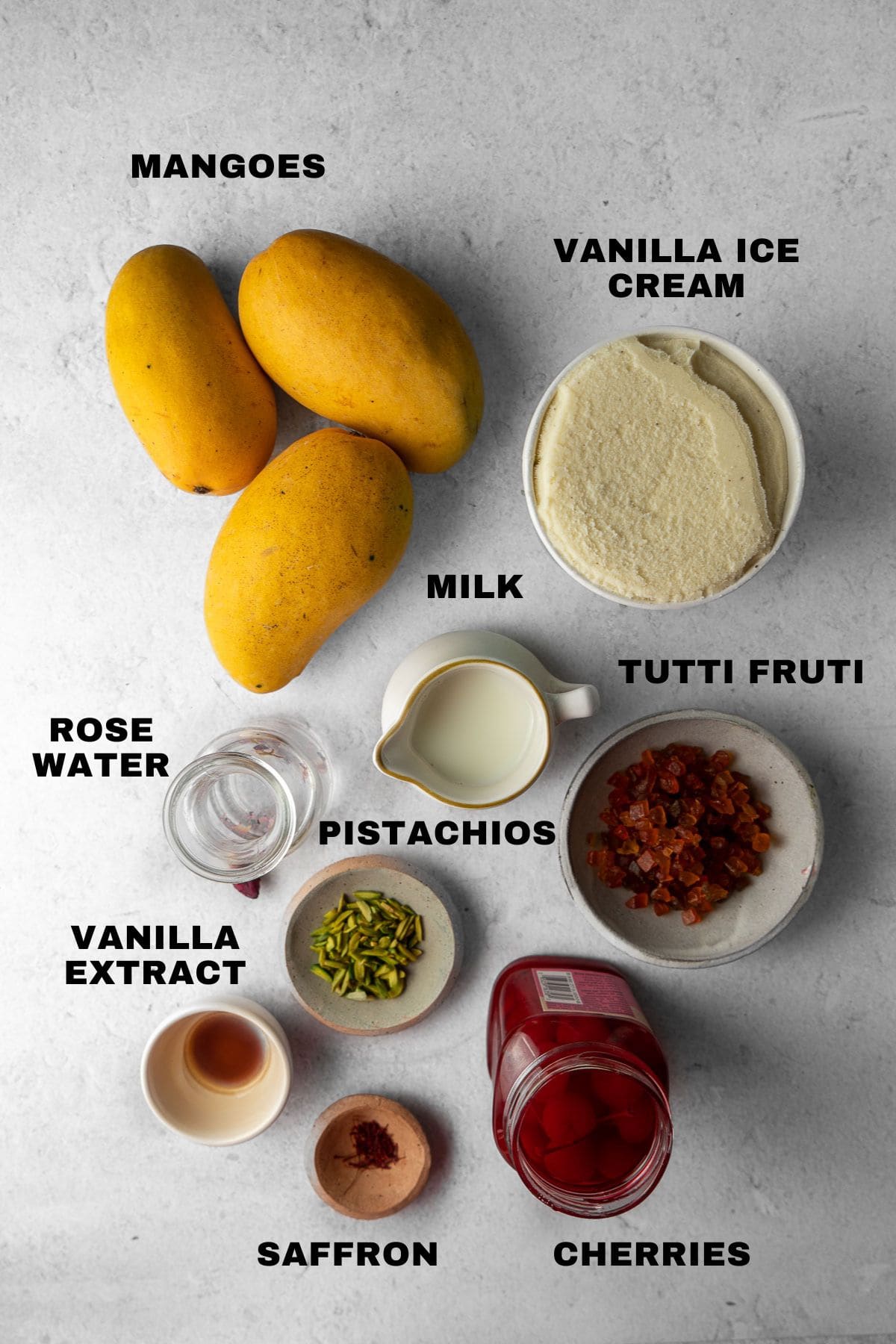 Ingredients for Mango Mastani with labels - mangoes, vanilla, milk, tutti frutti, pistachios, vanilla extract, saffron, cherries, rose water.