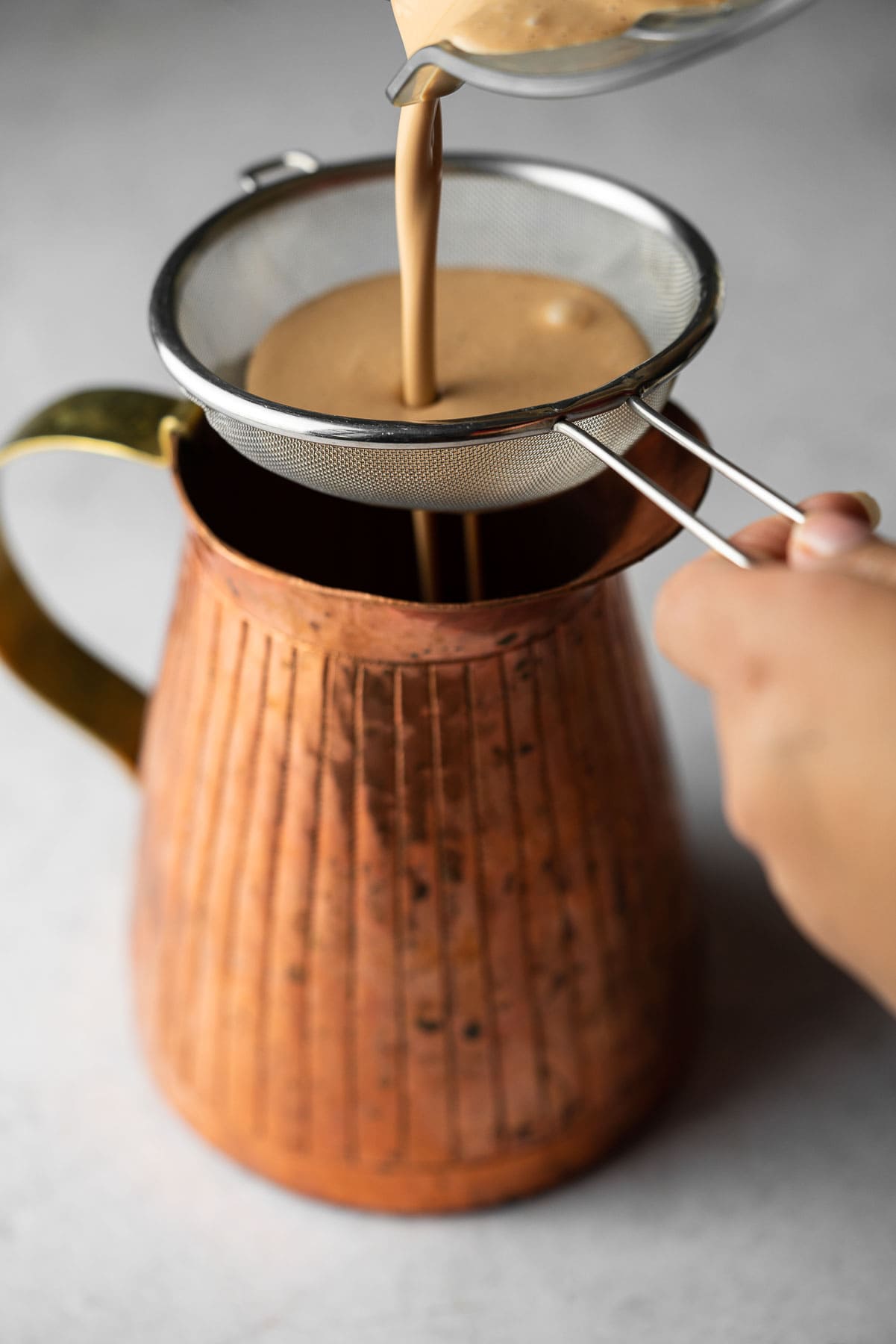 Straining chai horchata paste through a filter into a copper pitcher.