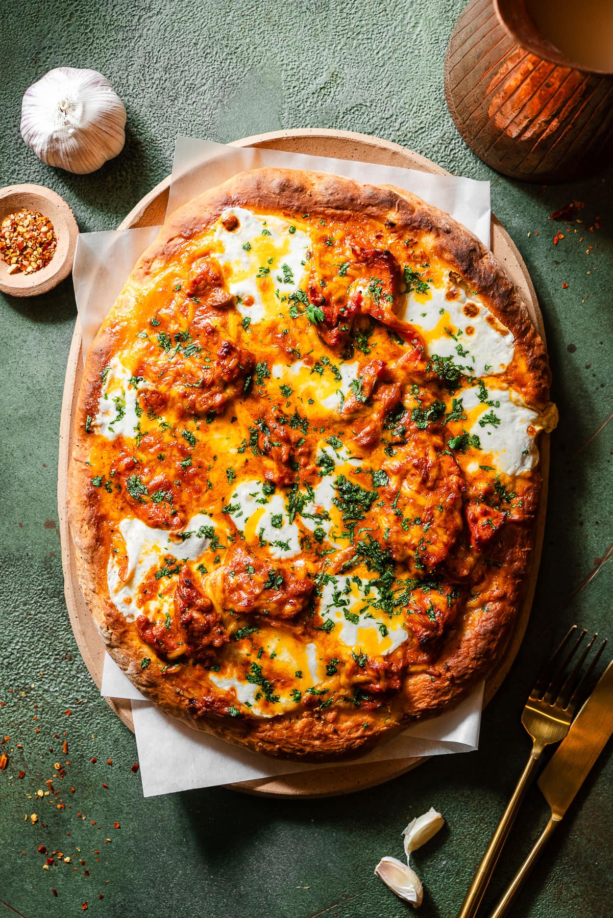 Butter Chicken Garlic Naan Pizza in a serving tray with cloves and a head of garlic, chili falkes, and a fork and knife.