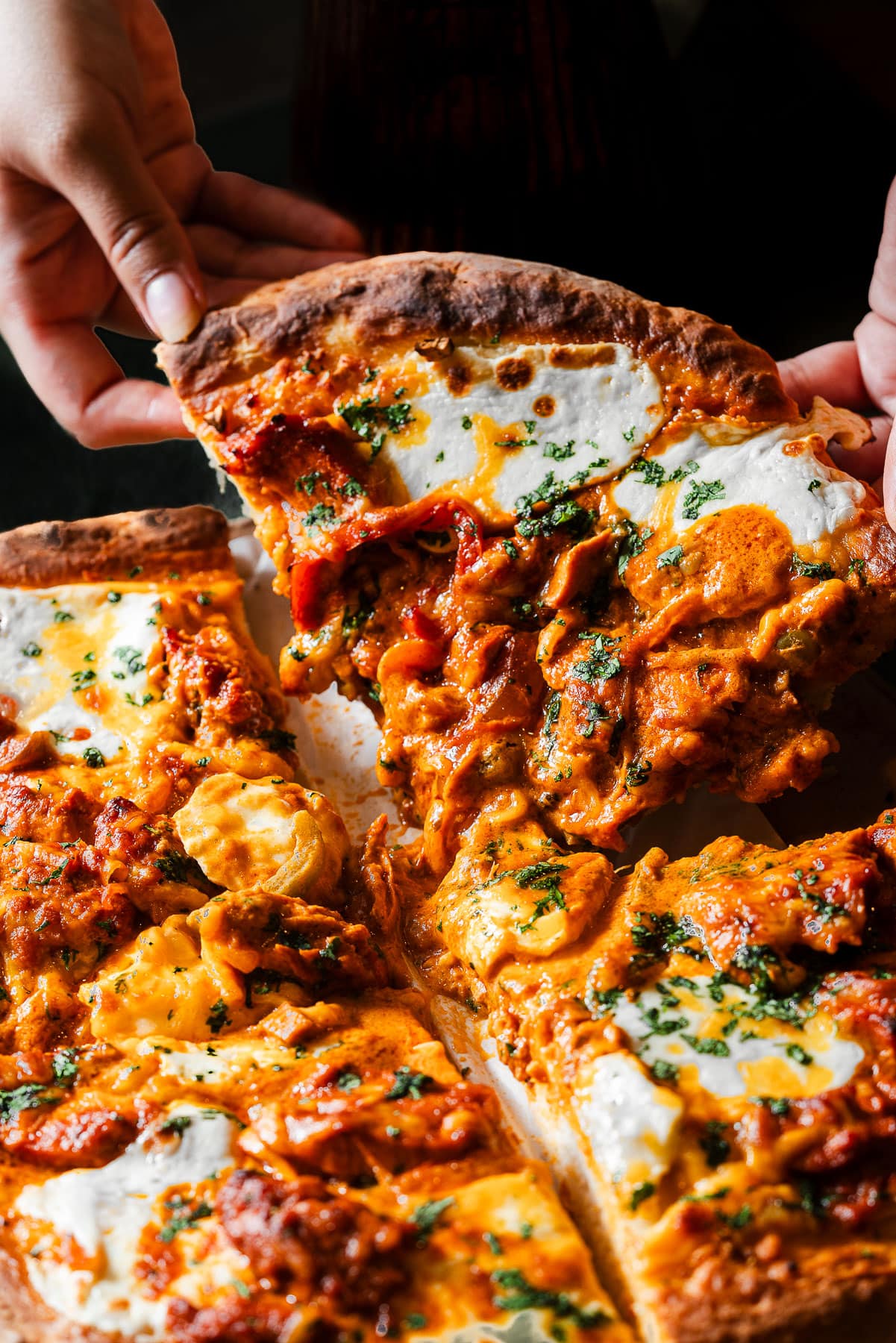 Two hands grabbing a large slice of garlic naan butter chicken pizza.