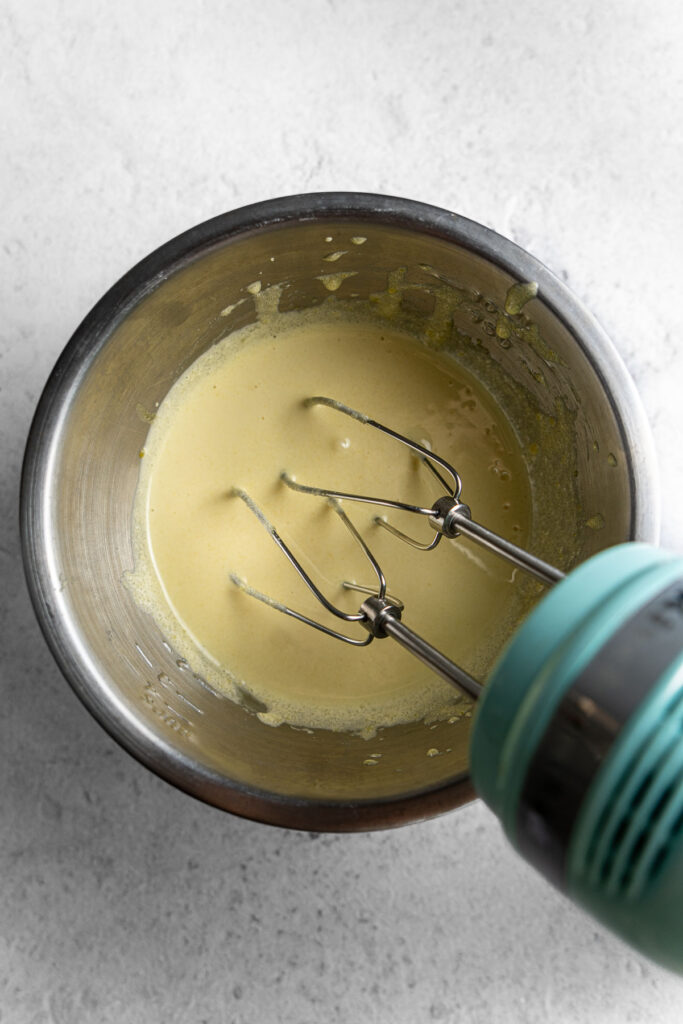 A handheld electric mixer beating egg yolks and sugar until pale yellow.