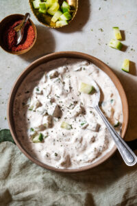 Cucumber Mint Raita in a bowl with a spoon with chili powder and extra cucumbers.