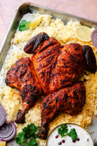Roasted Spatchcock Tandoori Chicken in a baking tray with rice, onion, lemons, and raita.