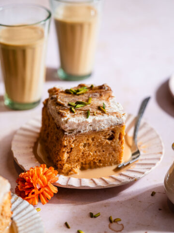 A slice of masala chai tres leches cake in a cake plate with two cups of masala chai.
