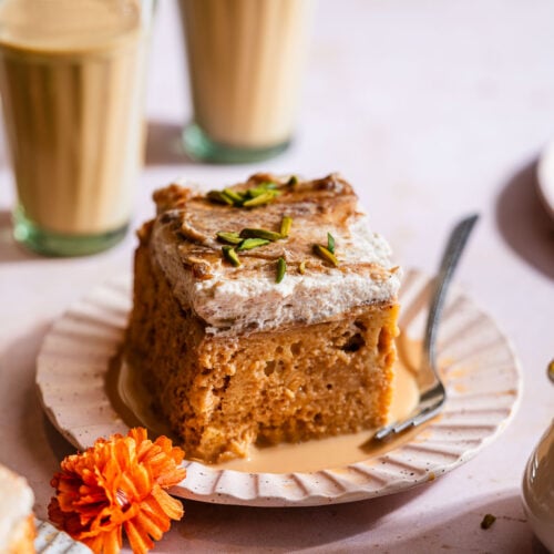 A slice of masala chai tres leches cake in a cake plate with two cups of masala chai.
