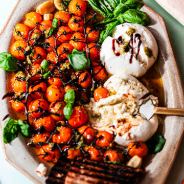 A platter full of roasted cherry tomatoes with grilled bread, basil, and two pieces of burrata.
