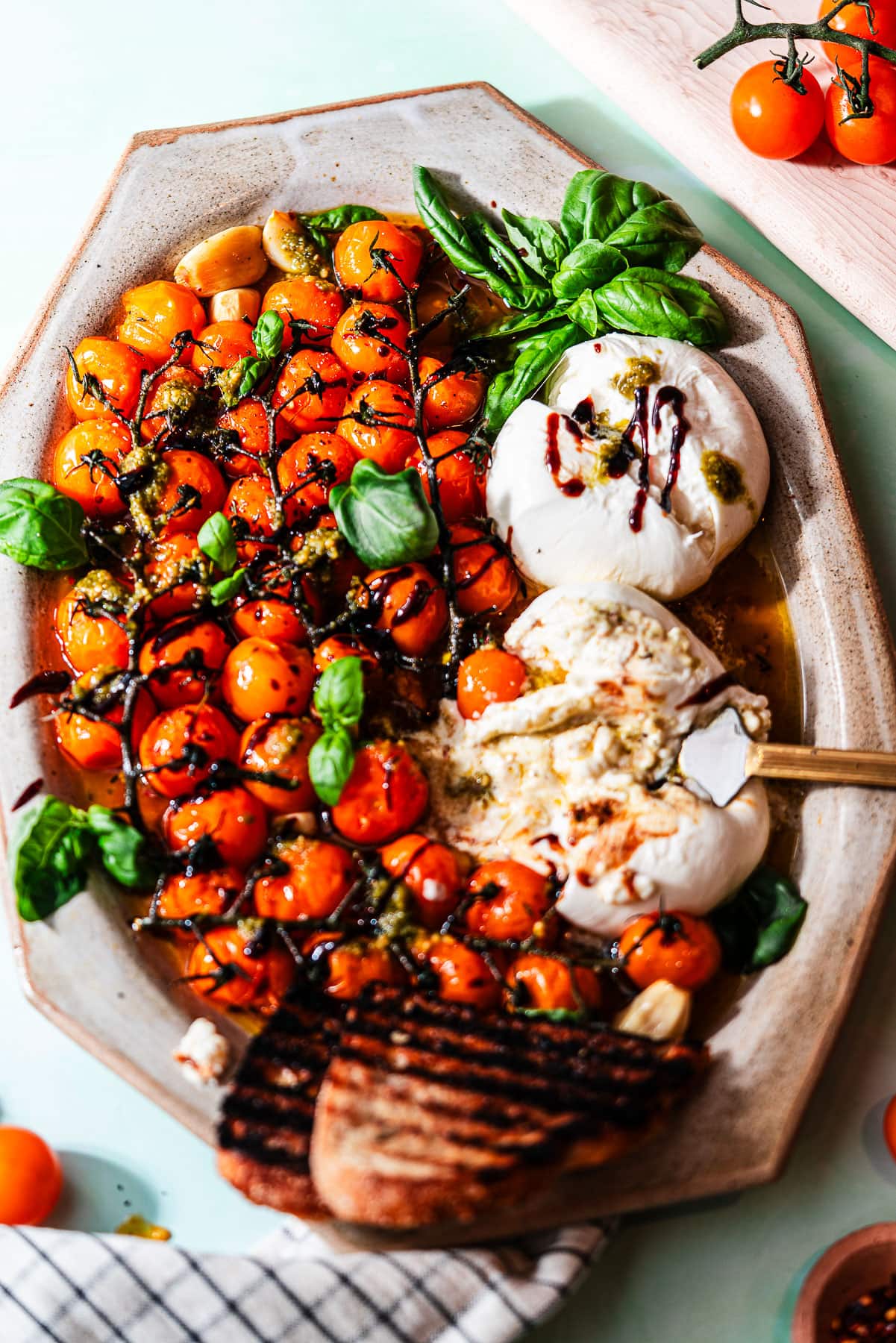 A platter full of roasted cherry tomatoes with grilled bread, basil, and two pieces of burrata.