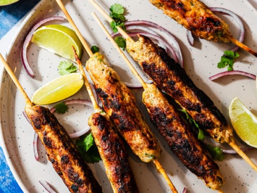 Chicken seekh kebabs on skewers in a plate with limes, onion, and cilantro.