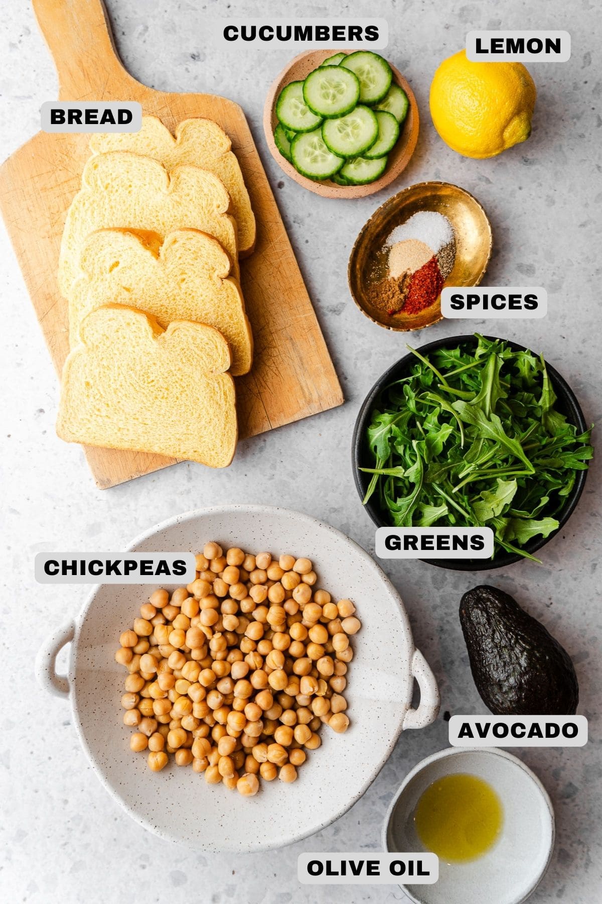 Bread, cucumbers, lemon, spices, greens, avocado, chickpeas, olive oil ingredients with labels.