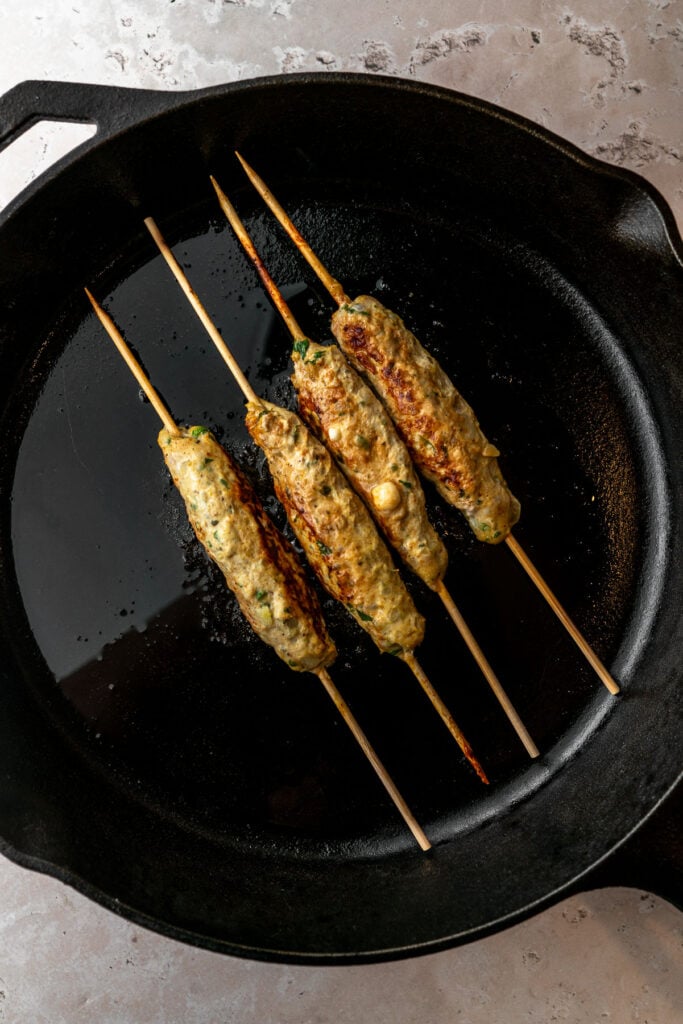 Cooking four chicken seekh kebabs on skewers in a cast iron pan.