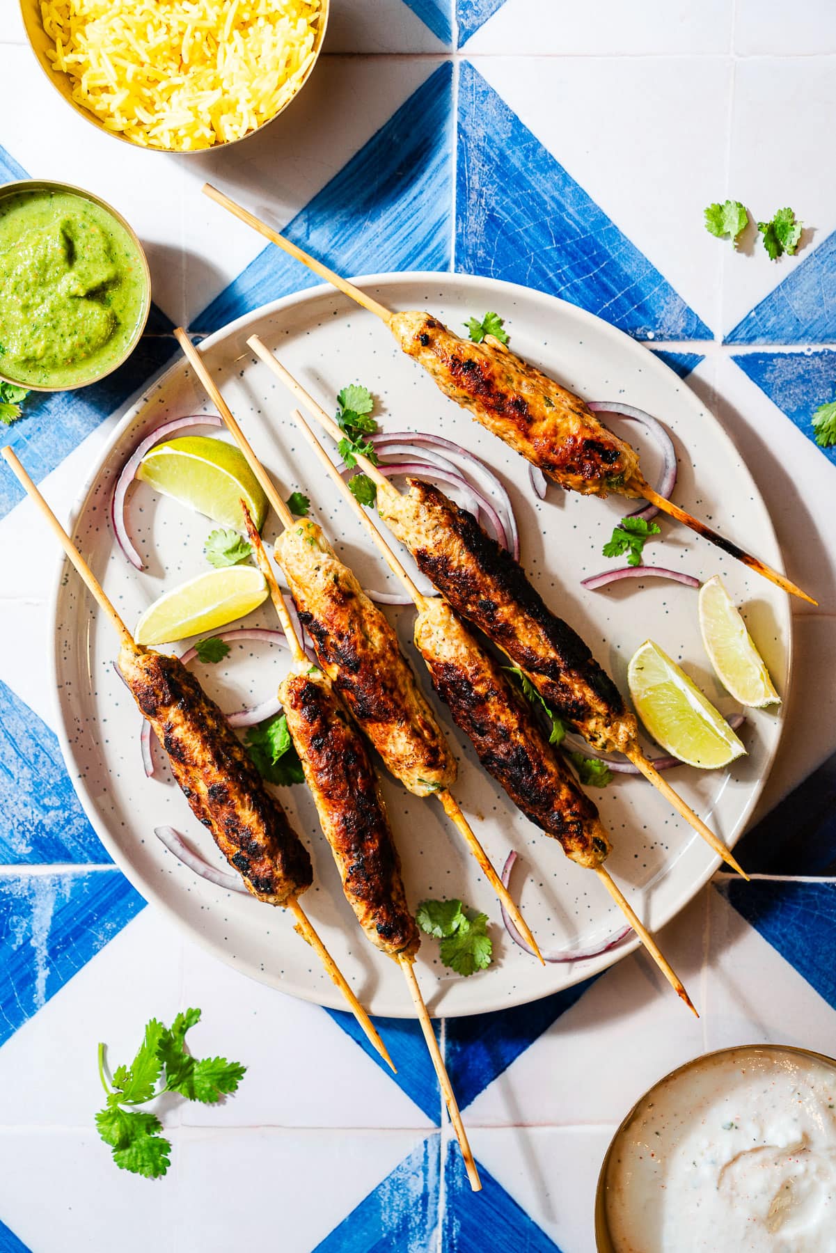 Chicken seekh kebabs on skewers in a plate with limes, onion, and cilantro with chutney, rice, and raita bowls.