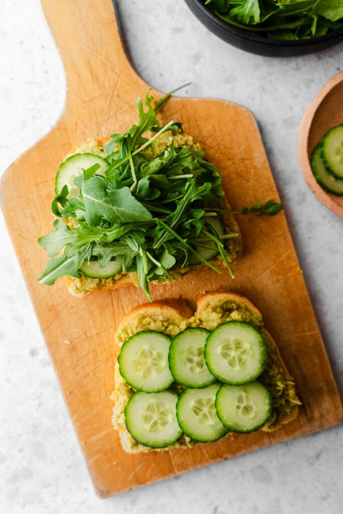 Cucumbers and baby arugula layering a chickpea avocado sandwich on a cutting board.