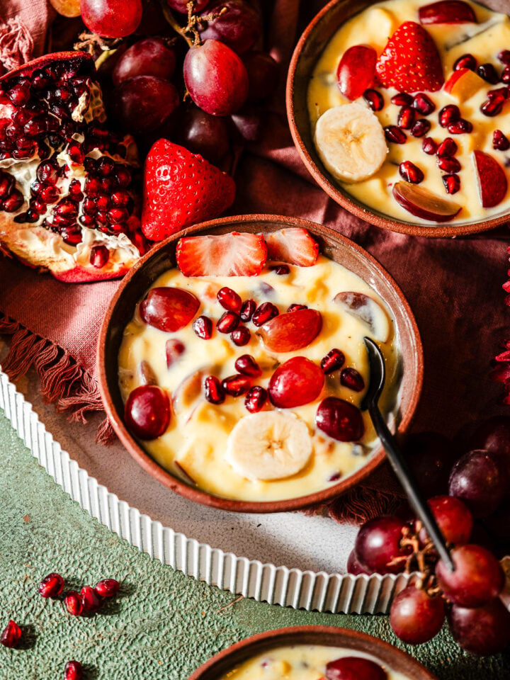 Three dessert bowls of fruit custard in a platter filled with fruits and flowers.