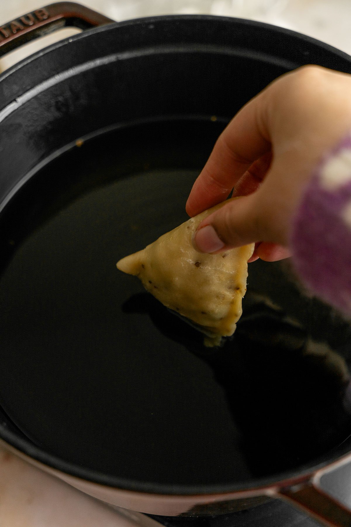 A hand dropping a chicken samosa into a pot of hot oil.
