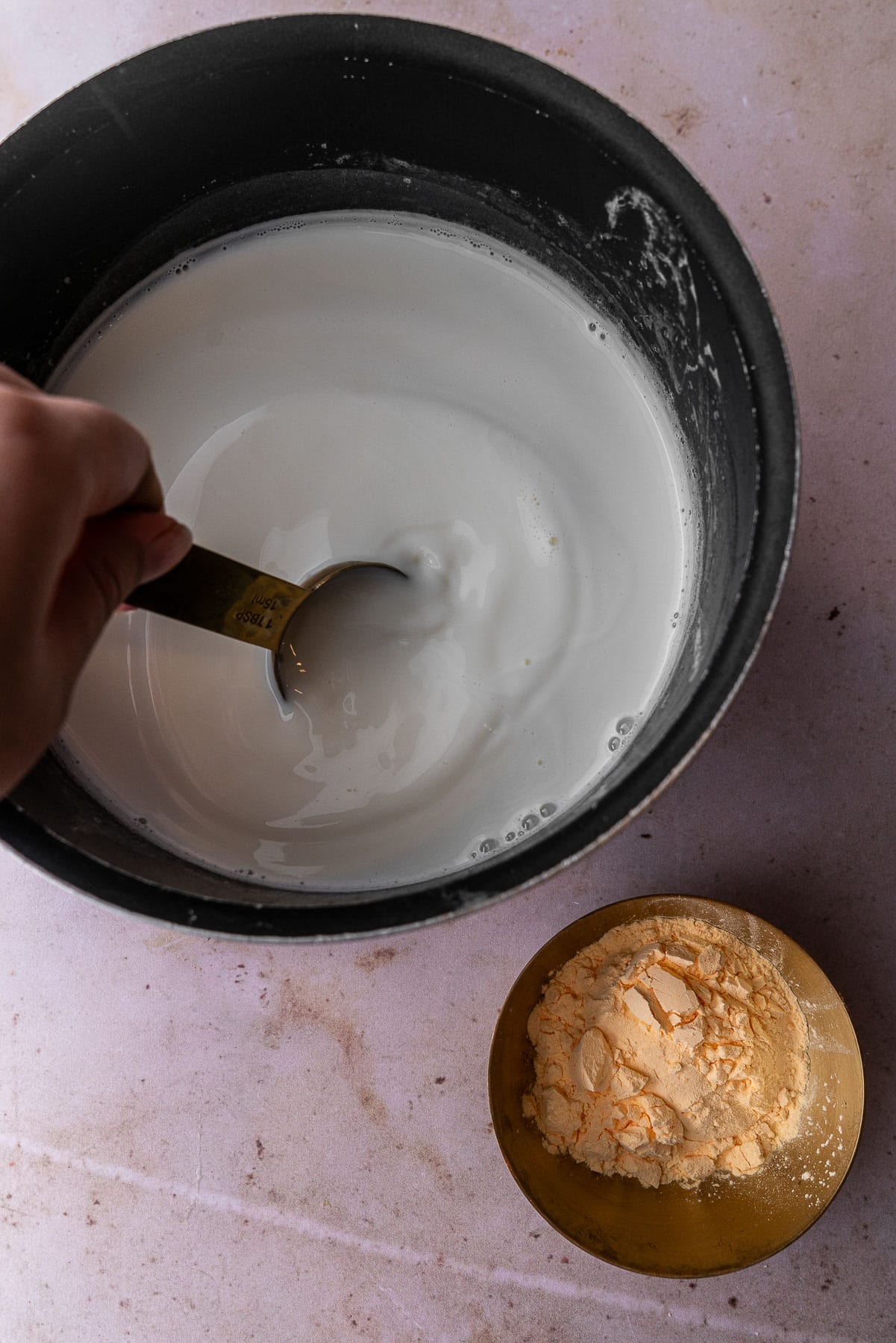A tablespoon scooping up milk in a pan to pour into a bowl with custard powder.