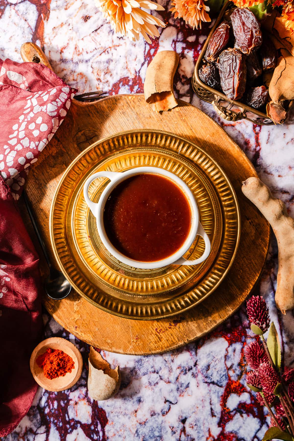 A cocotte full of imli chutney in layered gold plates surrounded by fresh tamarind, dates, and flowers.